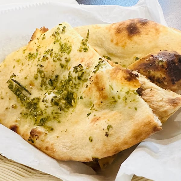 Close-up of golden-brown Pakistani naan bread, topped with vibrant green herbs, served in a white paper-lined basket.