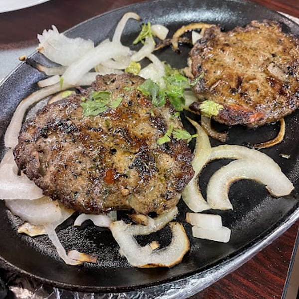 Two sizzling Pakistani chapli kababs with grilled onions and cilantro on a black platter.